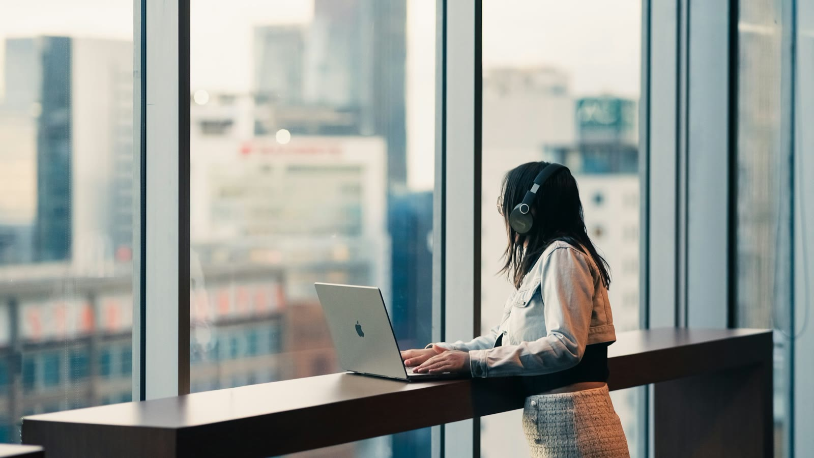 A woman working at her laptop by a city window, thinking about her career